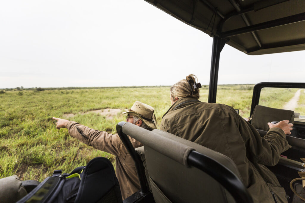 Adult woman and a safari guide leaning out looking across a plain to animals in the distance.